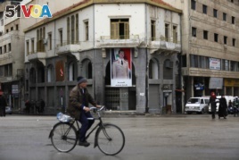 A man rides his bicycle past by a banner showing Syrian President Bashar Assad, at the clock square in the old city of Homs, Syria, Jan. 17, 2018.