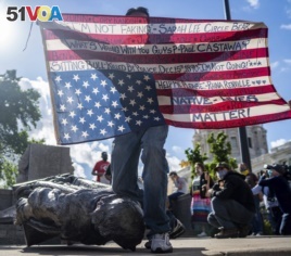 Gabriel Black Elk, who is Lakota, kneels on the neck of a fallen statue of Christopher Columbus and holds an American flag with the names of Native Americans killed by police, at the Minnesota state Capitol in St. Paul, Minn., June 10, 2020. (Evan Frost)